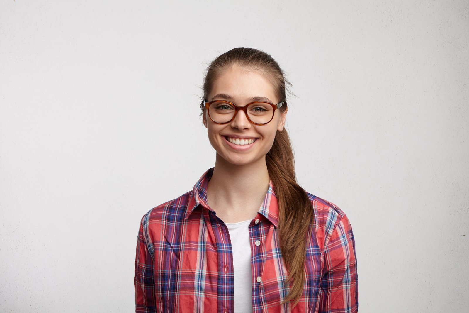 Headshot of attractive teenage girl with cute nice features and ponytail looking at camera and smiling cheerfully, enjoying good day and happy moments of her youth, dressed in stylish clothing